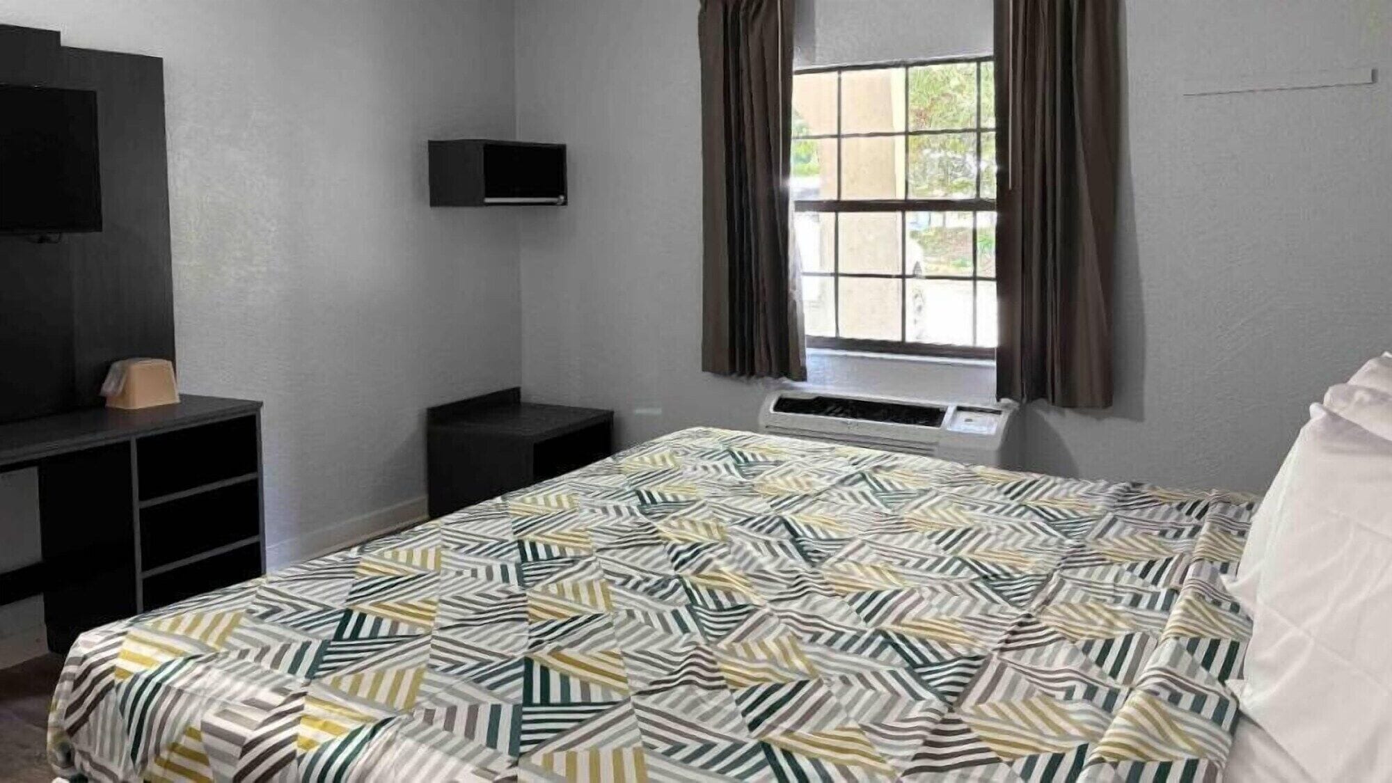 A bedroom with a bed, a TV mounted on the wall, a window with curtains, and a heater.