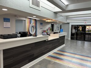 A reception desk with a large number '6' sign, a computer, and a printer. There is a sign for Studio 6 amenities and an exit sign. - Studio 6 Tallahassee, FL – North Monroe (Tallahassee)