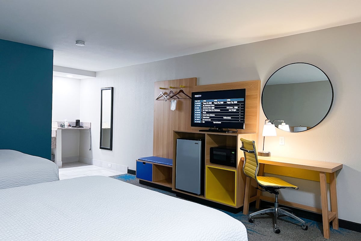 A hotel room with a bed, a desk with a computer, a chair, a small fridge, a microwave, and a TV displaying a menu.