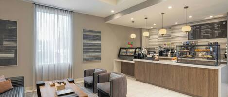 A modern hotel lobby with a seating area, a coffee bar, and a large window with curtains.