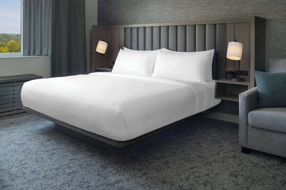 A modern hotel room with a large bed, two bedside tables with lamps, a grey upholstered armchair, and a view of the outdoors through a window.