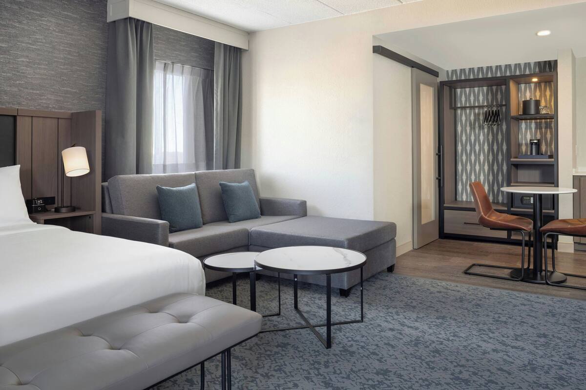 A modern hotel room with a large bed, a gray sofa with blue cushions, two small round tables, and a dining area with a chair and table.