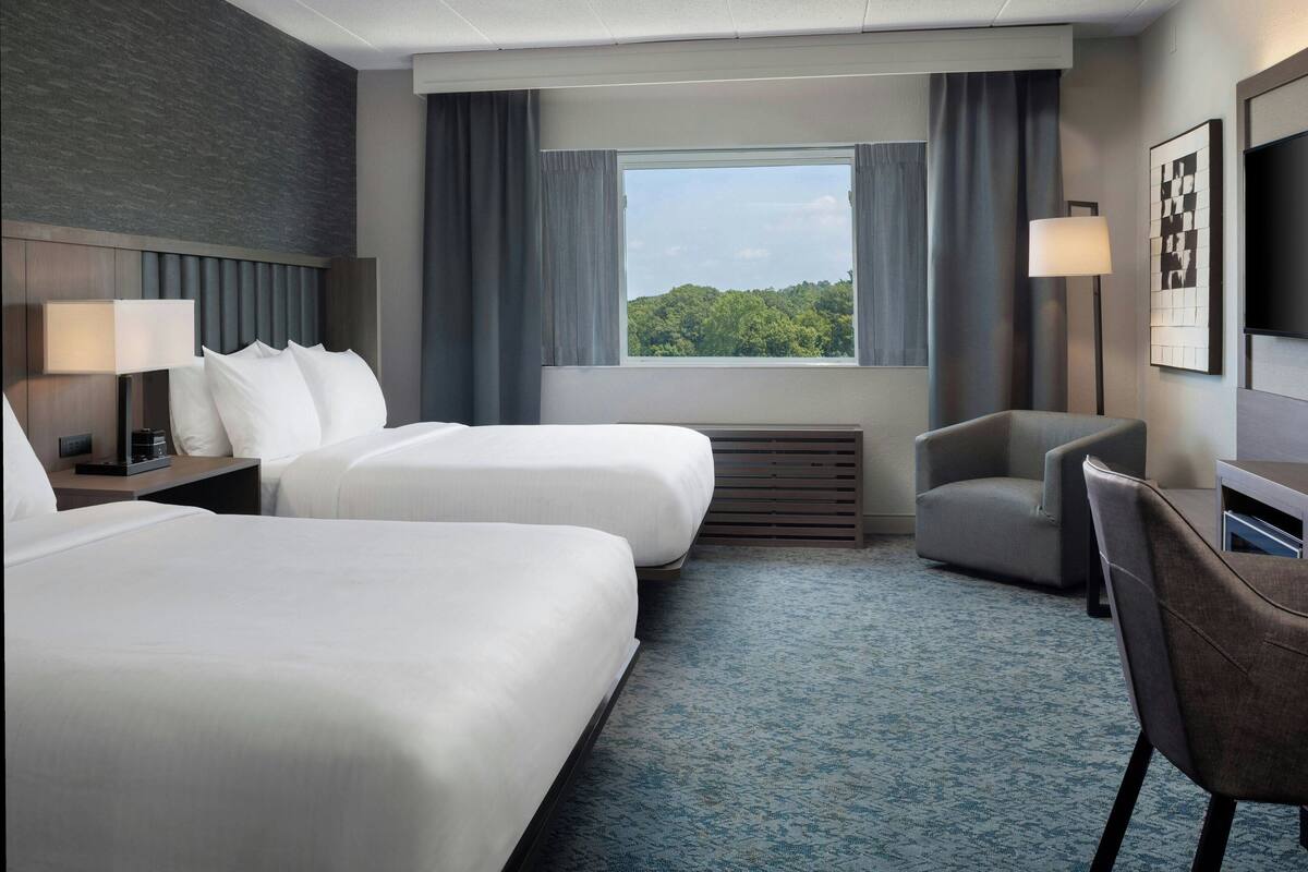 A hotel room with two beds, a desk, a chair, a TV, and a window with a view of greenery.