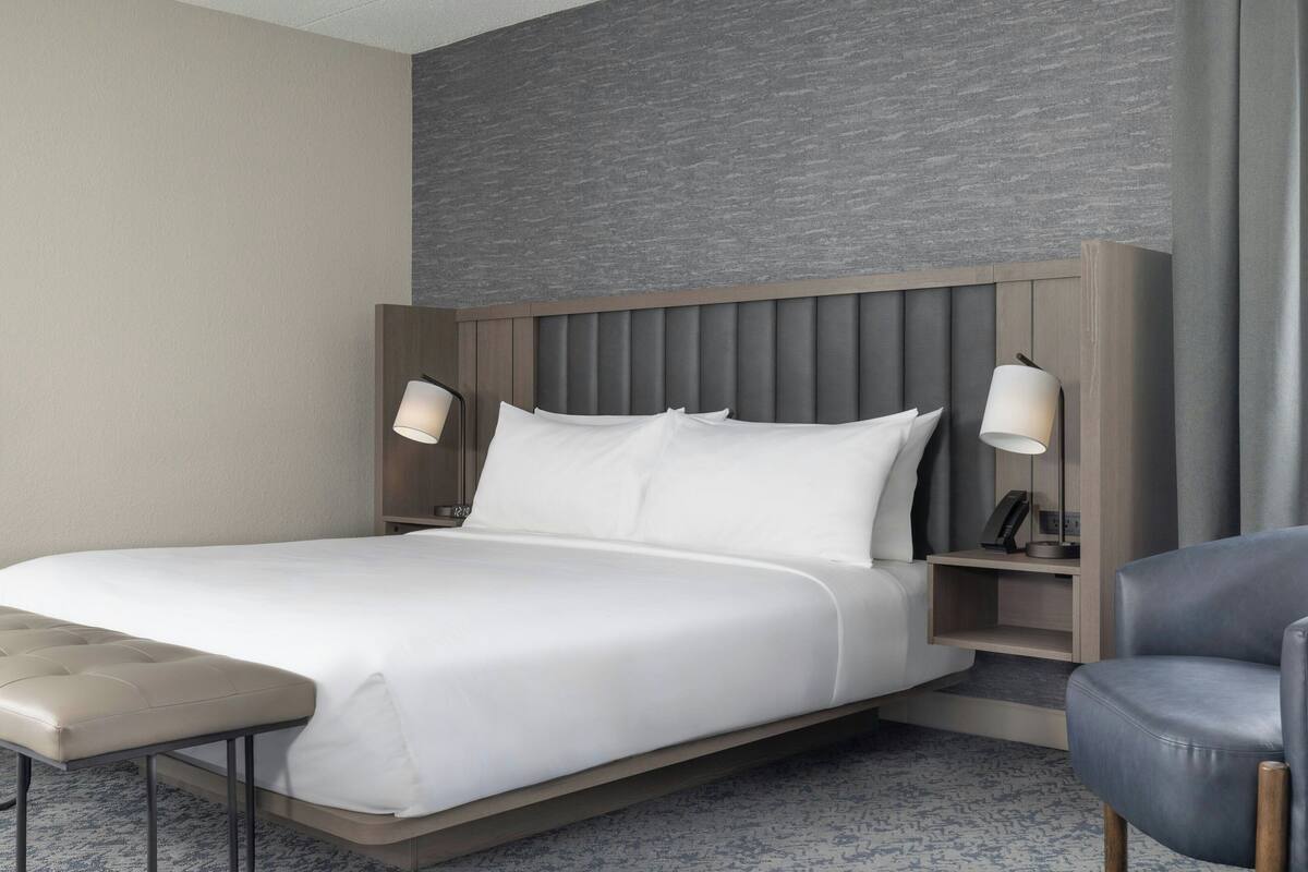 A modern hotel room with a large bed, a bench, two bedside tables with lamps, and a grey textured wall.