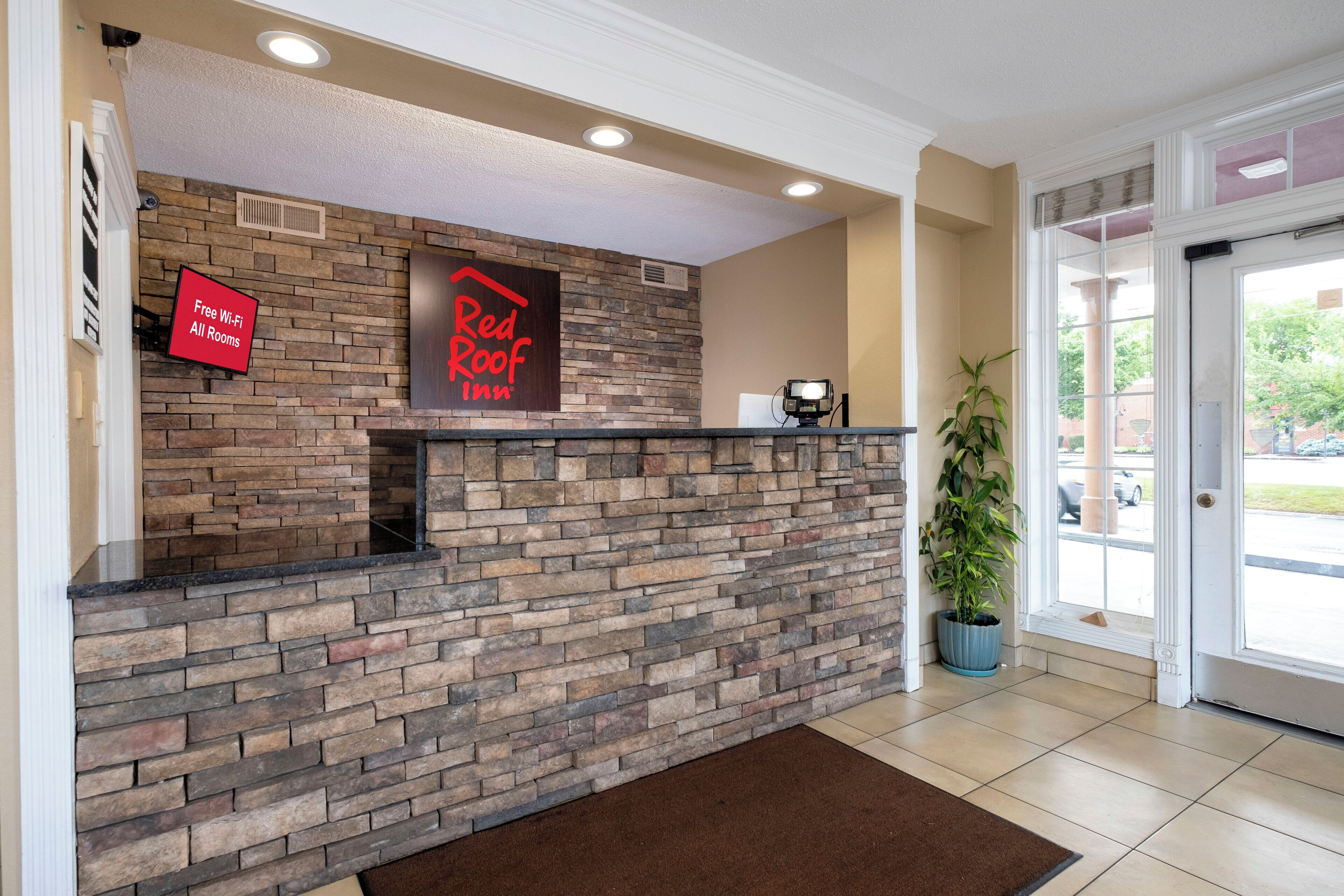 A reception desk with a stone wall, a Red Roof Inn sign, a 'Free Wi-Fi All Rooms' sign, and a potted plant.