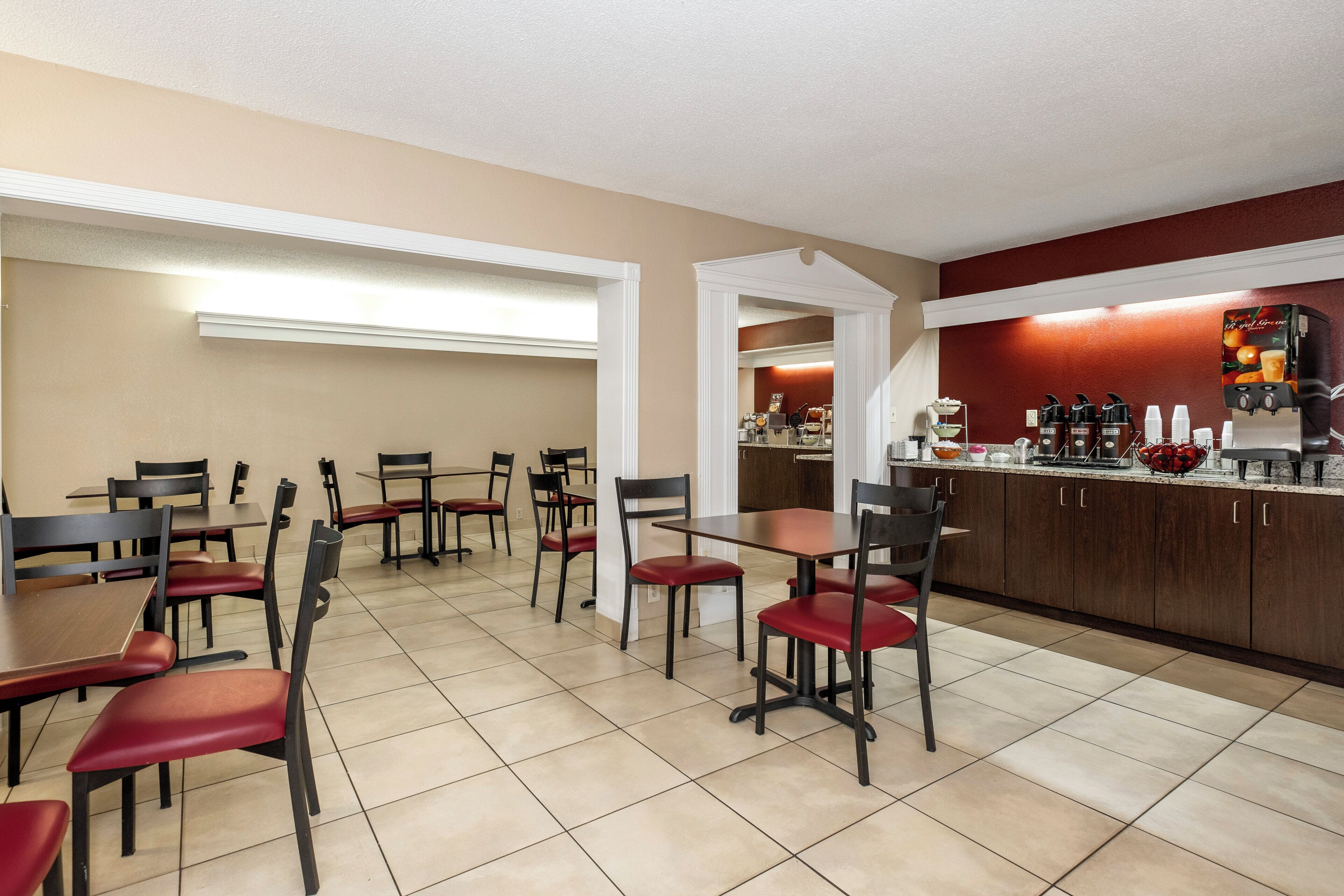 Un comedor de hotel con mesas y sillas, una zona de cocina con cafeteras y un mostrador con dispensador de bebidas.