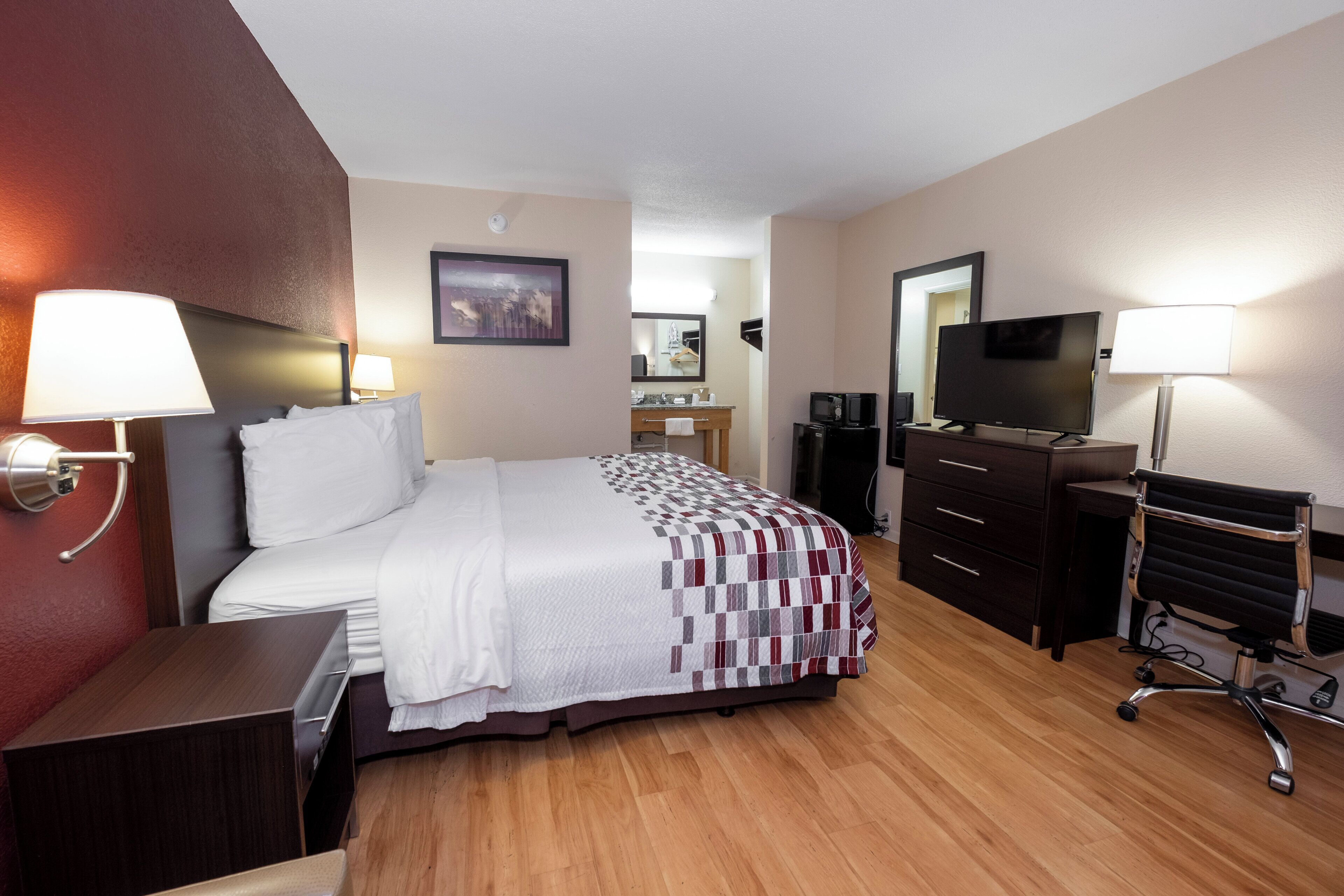 Una habitación de hotel con cama, mesitas de noche, escritorio, silla, televisor y espejo.