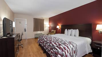 A hotel room with a bed, a desk with a computer, a chair, a TV, and a wall-mounted lamp.