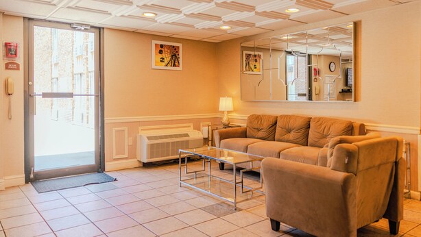 Een hotellobby met een bank, een glazen bijzettafeltje, een radiator, een deur met spiegel en een hangklok.