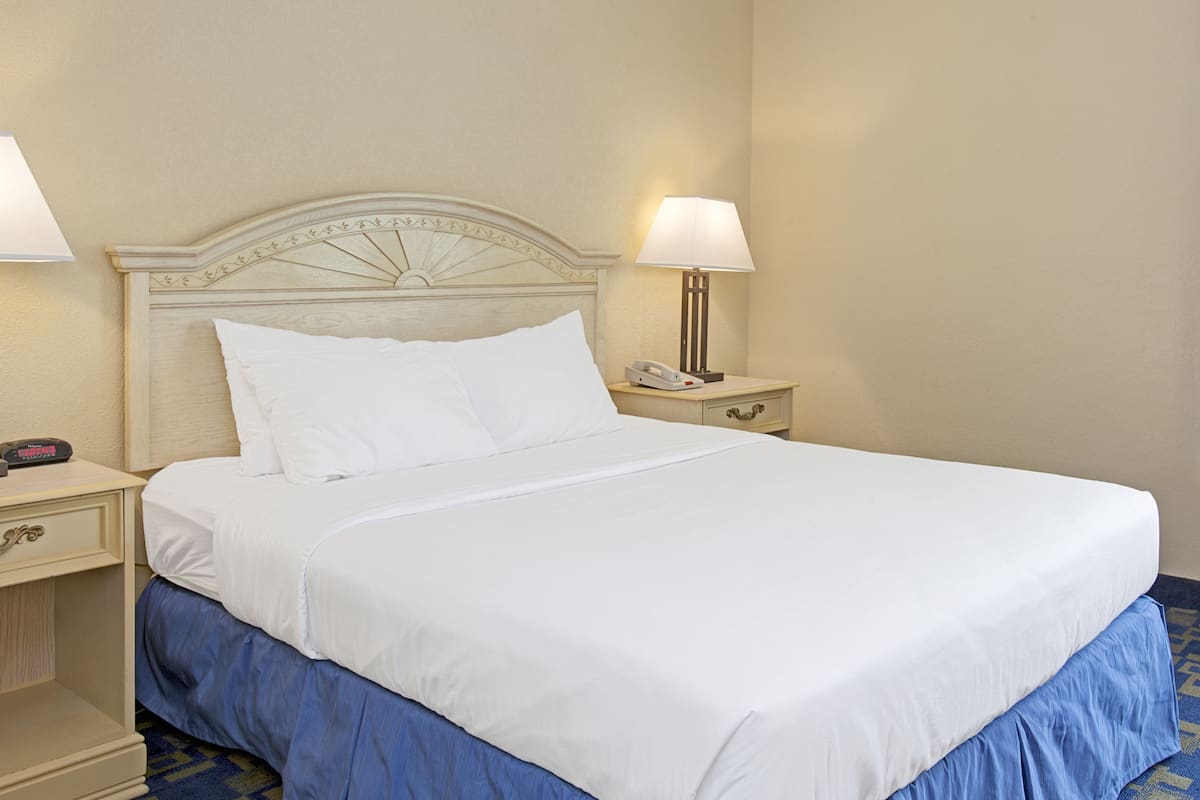 A neatly made bed with white linens and a blue skirt, a bedside lamp, and a nightstand with a phone.