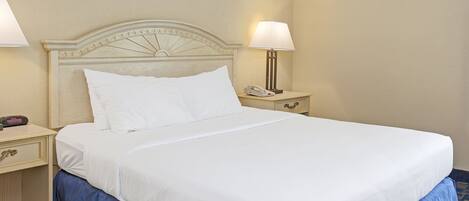 A neatly made bed with white linens and a blue skirt, a bedside lamp, and a nightstand with a phone.