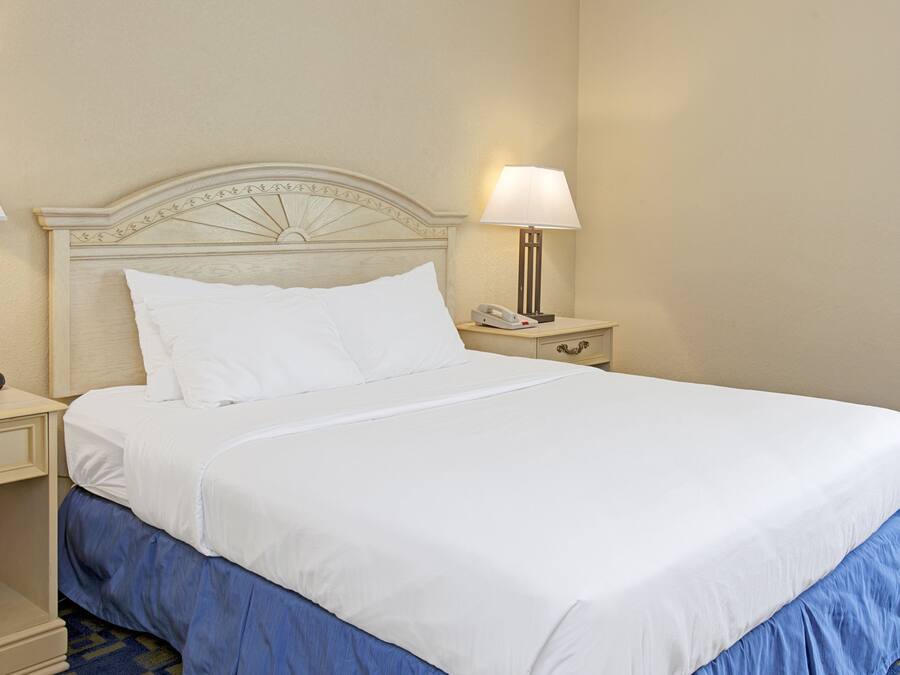 Una cama bien tendida con sábanas blancas y falda azul, una lámpara de noche y una mesita con un teléfono.