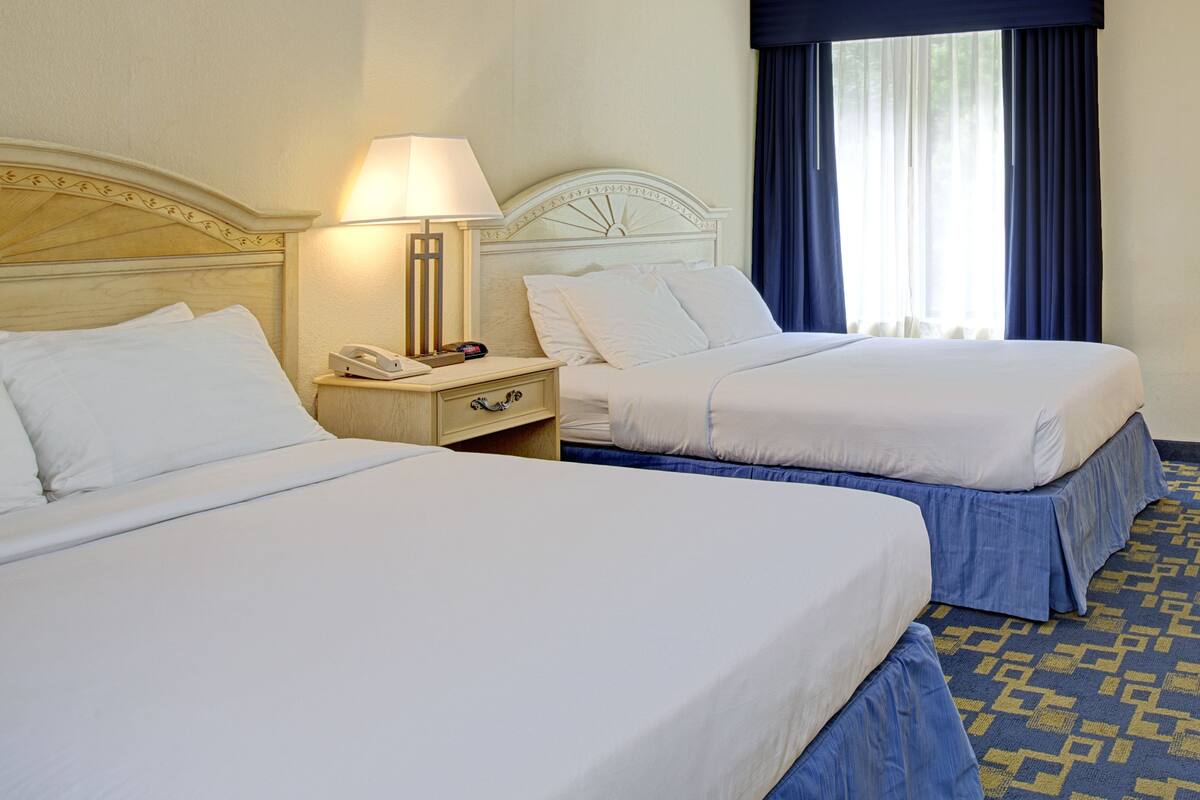 A hotel room with two beds, a nightstand with a lamp, and a window with curtains.