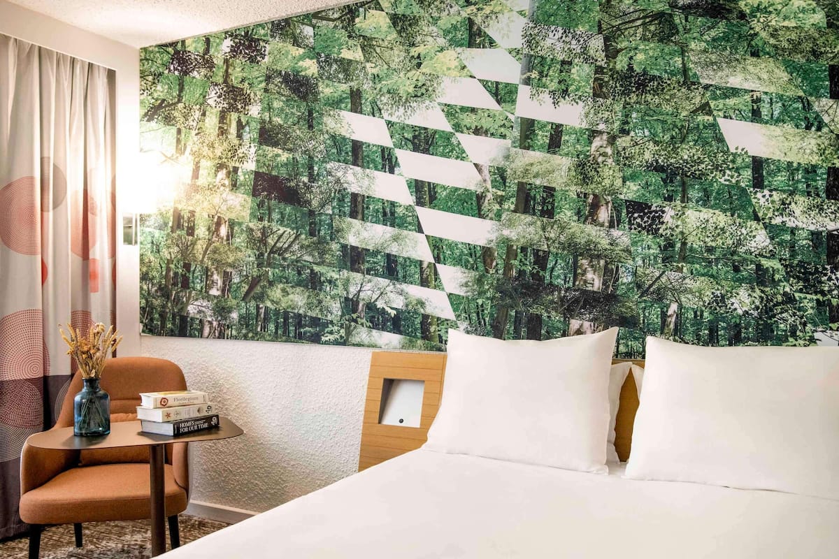 A hotel room with a bed, a chair, a small table with books, and a wall with a geometric forest pattern.