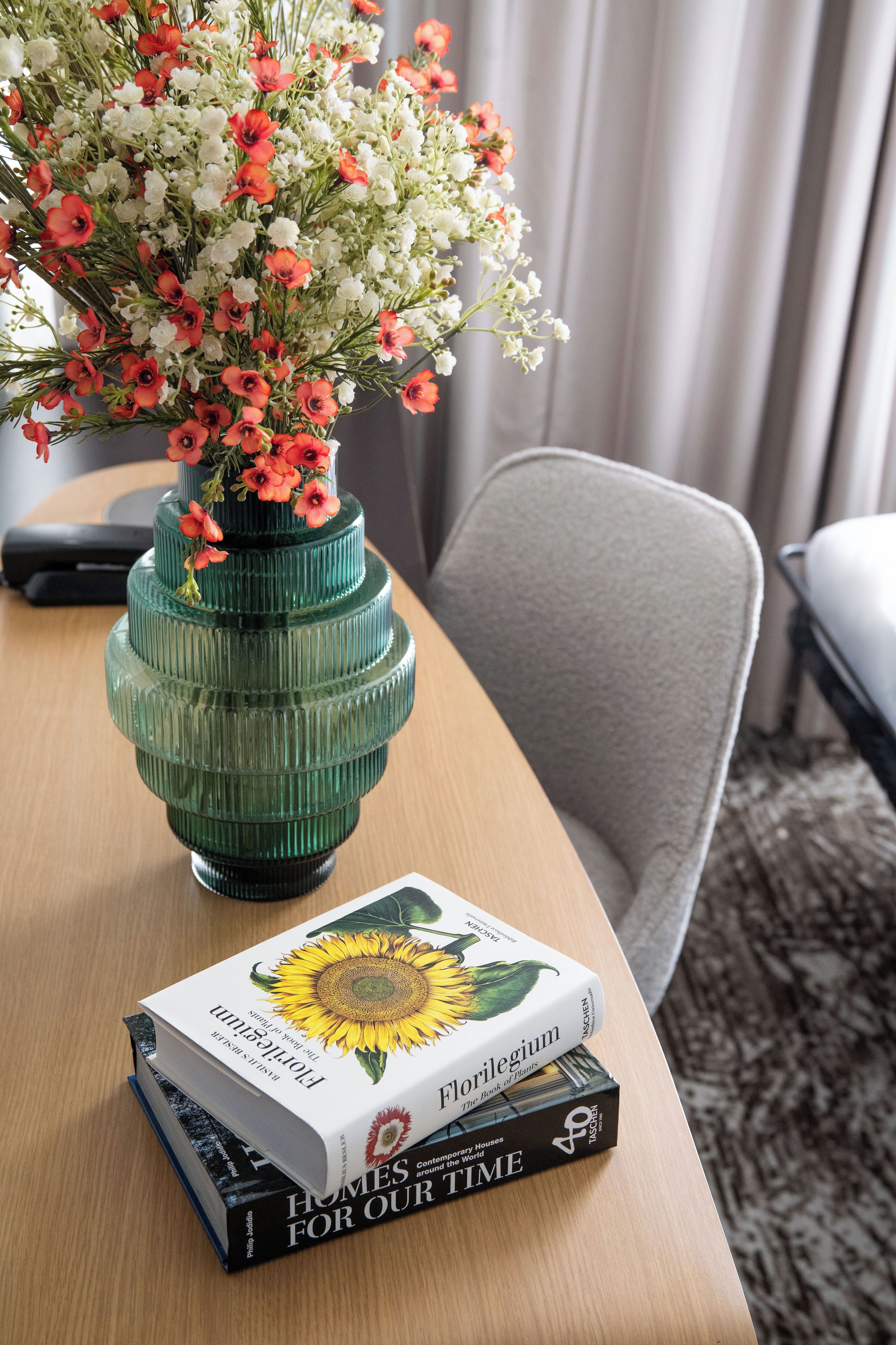Un vase vert orné de fleurs, posé sur une table en bois, à côté d’un livre intitulé « Des maisons pour notre époque ».