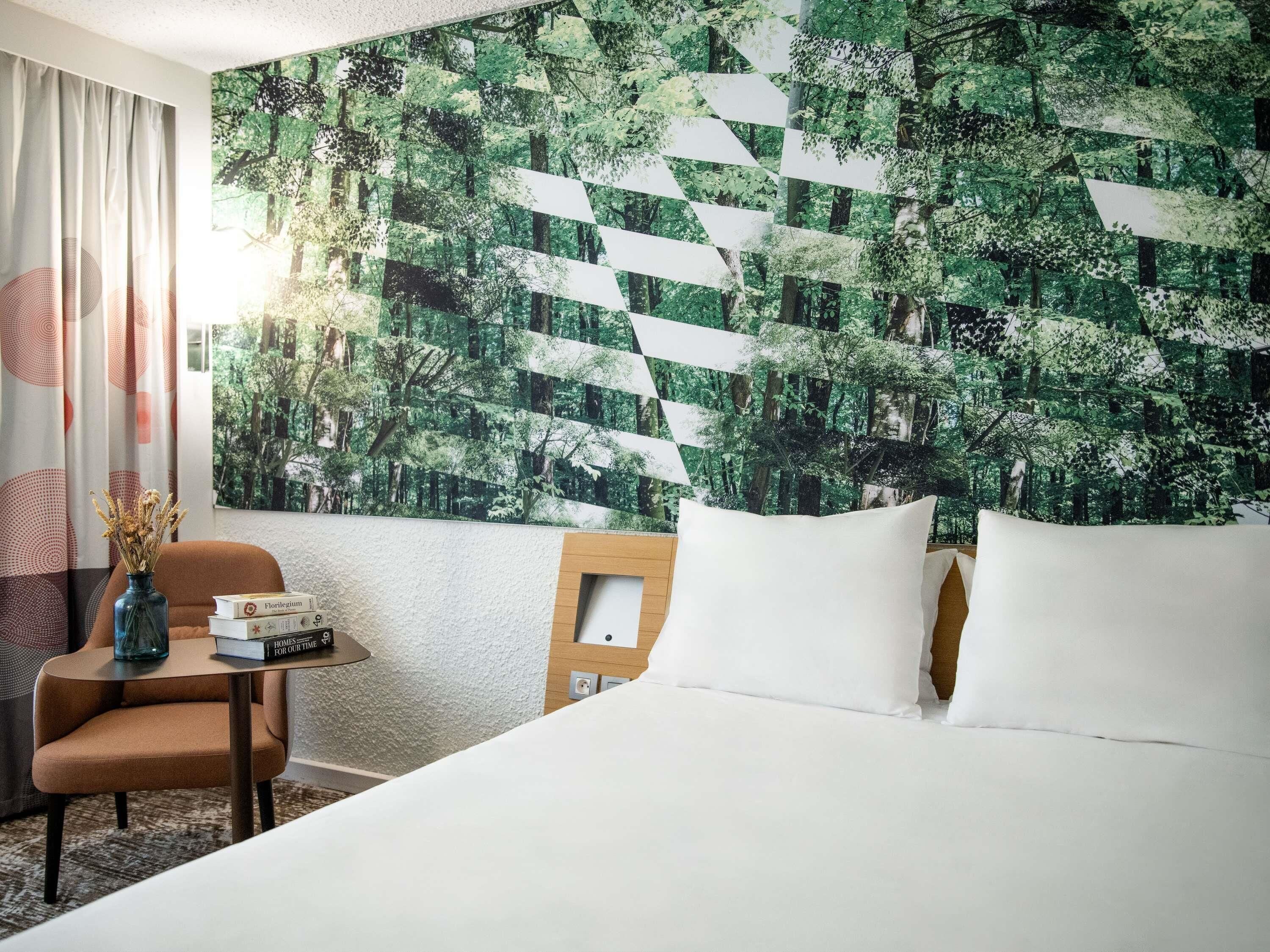 A hotel room with a bed, a chair, a small table with books, and a wall with a forest pattern.
