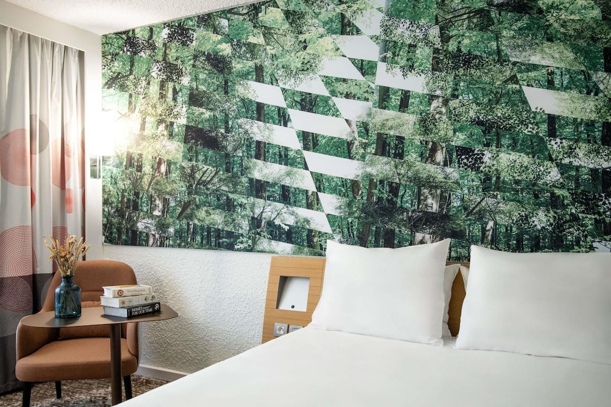 A hotel room with a bed, a chair, a small table with books, and a wall with a forest pattern.