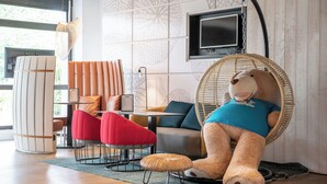 A modern interior with a large teddy bear in a wicker chair, red and gray chairs, a wooden table, and a TV on the wall.