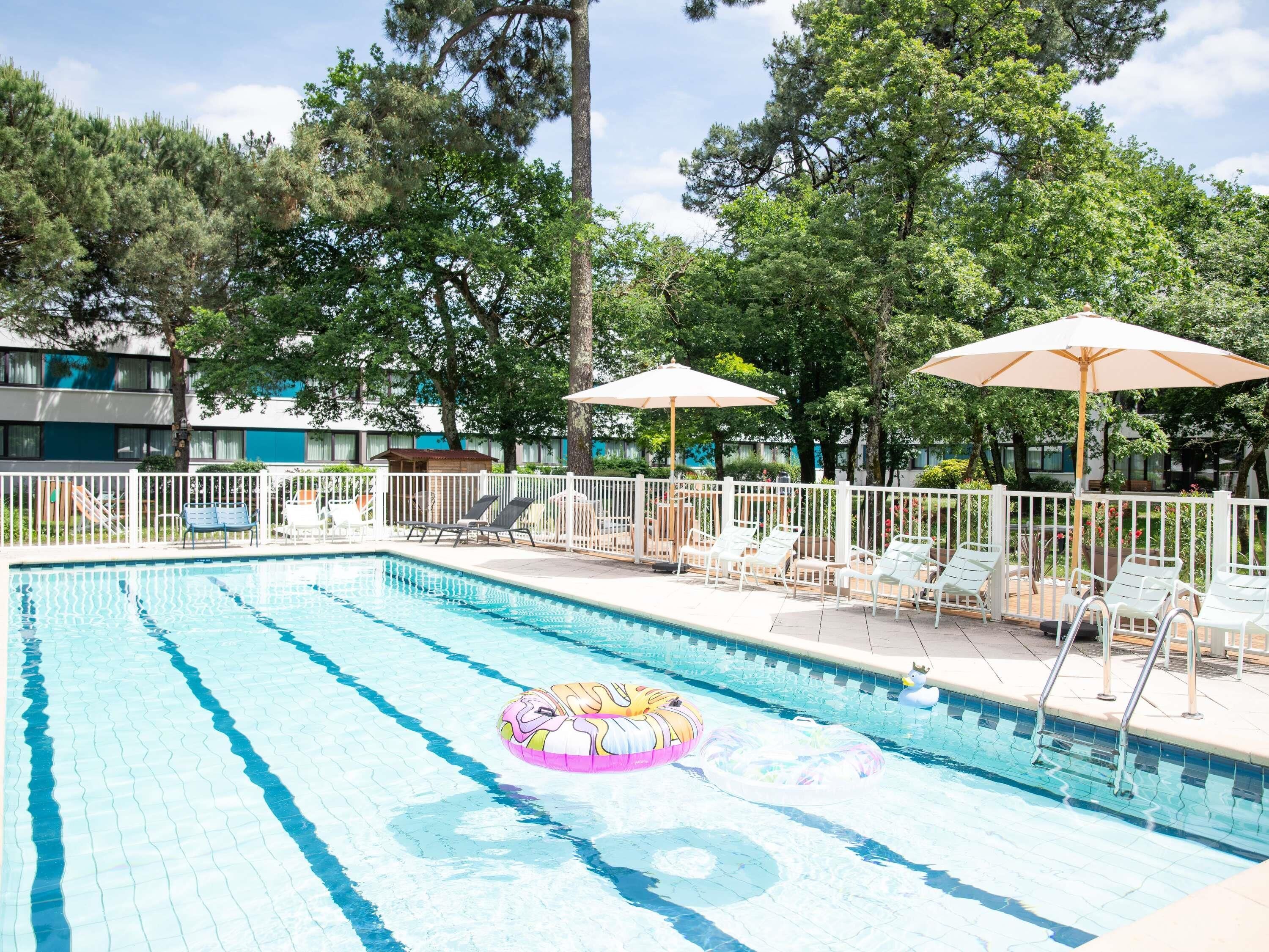 Une piscine entourée d’arbres et d’une clôture, avec des chaises longues et des parasols.
