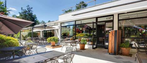 An outdoor patio area with tables and chairs, potted plants, and a glass storefront.