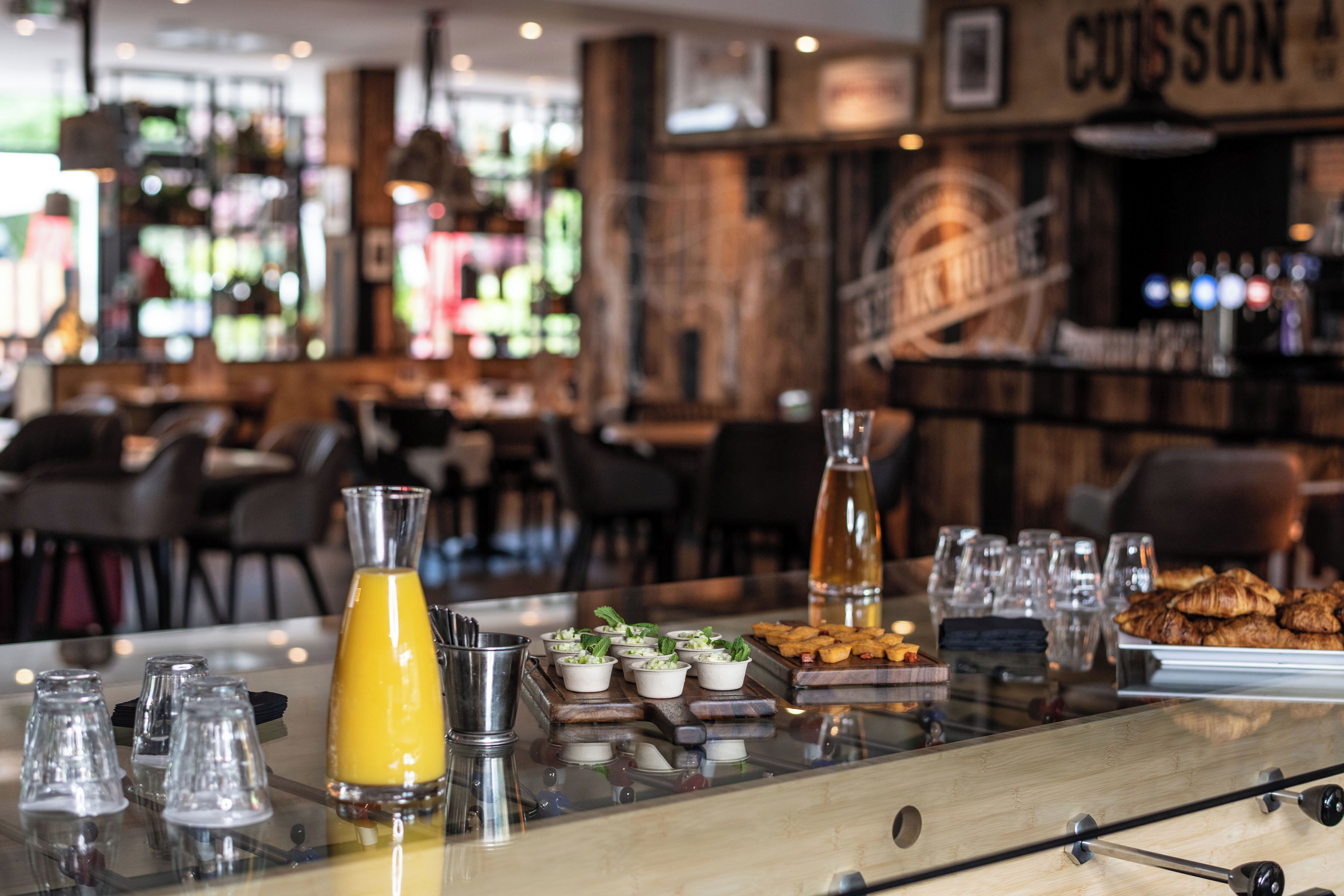 Un espace bar avec un comptoir en verre, des verres, une bouteille de jus d’orange et des produits alimentaires.