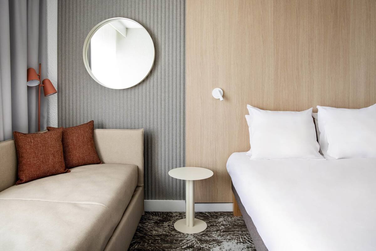 A hotel room with a bed, a sofa, a round mirror, a small table, and a lamp.