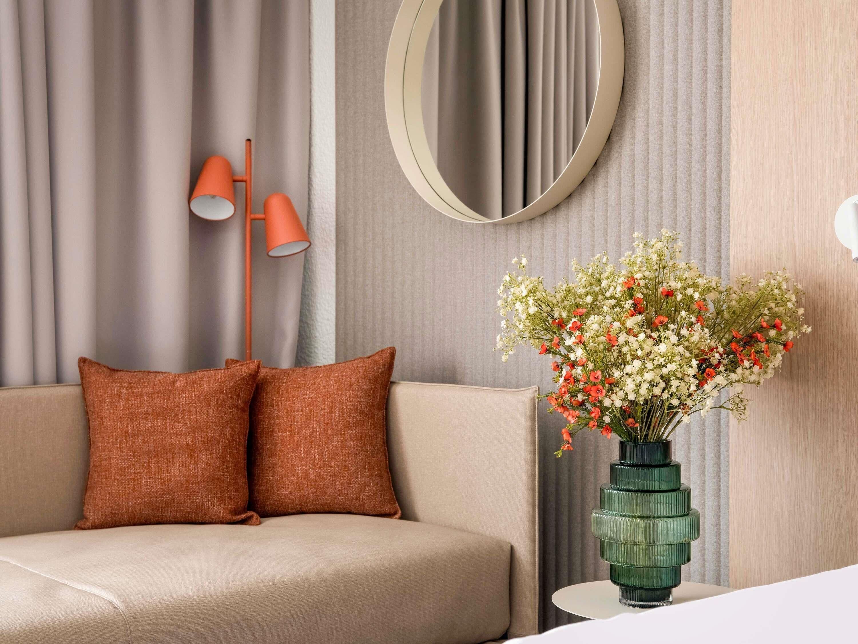 A modern living room with a sofa, two orange cushions, a green vase with flowers, a round mirror, and a floor lamp.