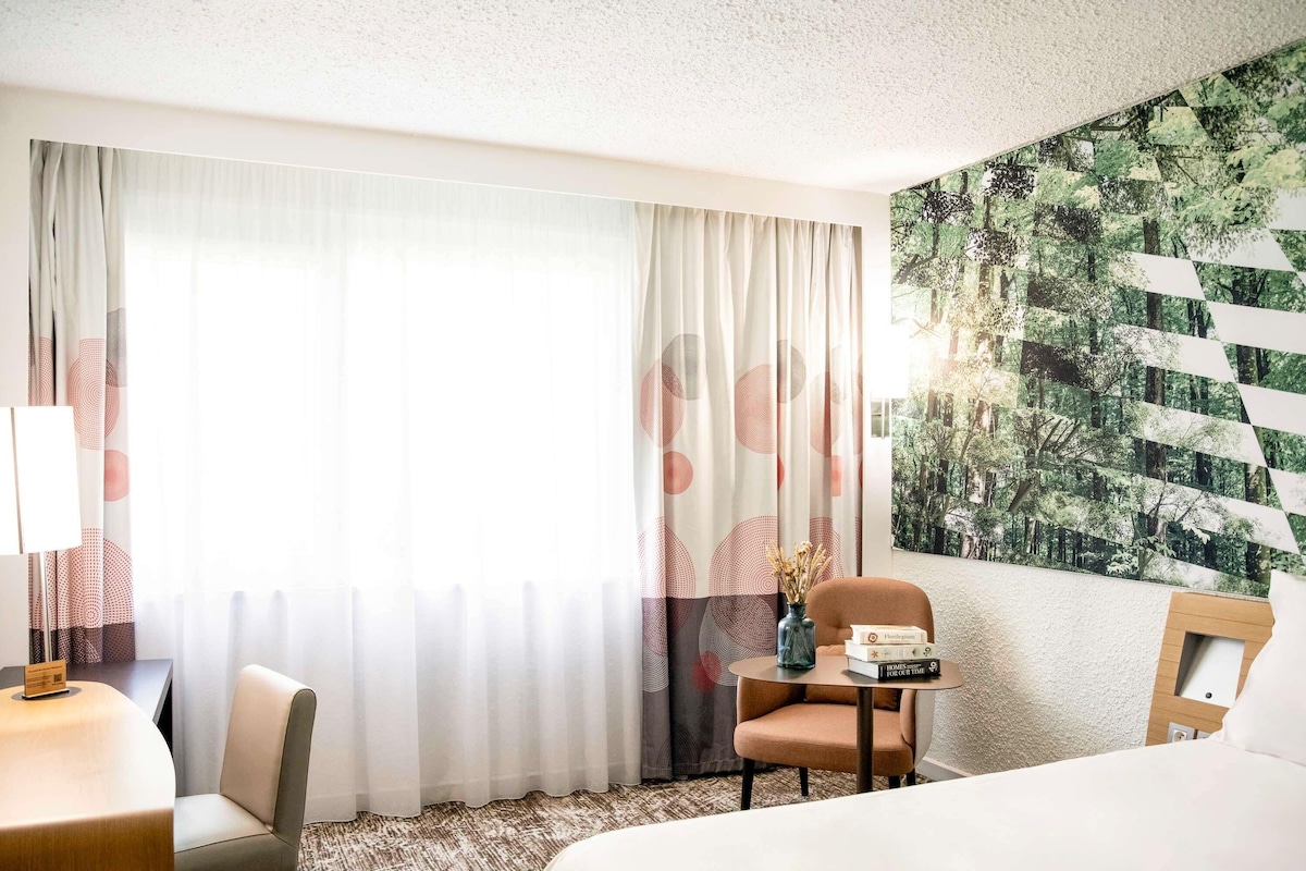 A hotel room with a bed, a desk with a lamp, a chair, and a small table with books.