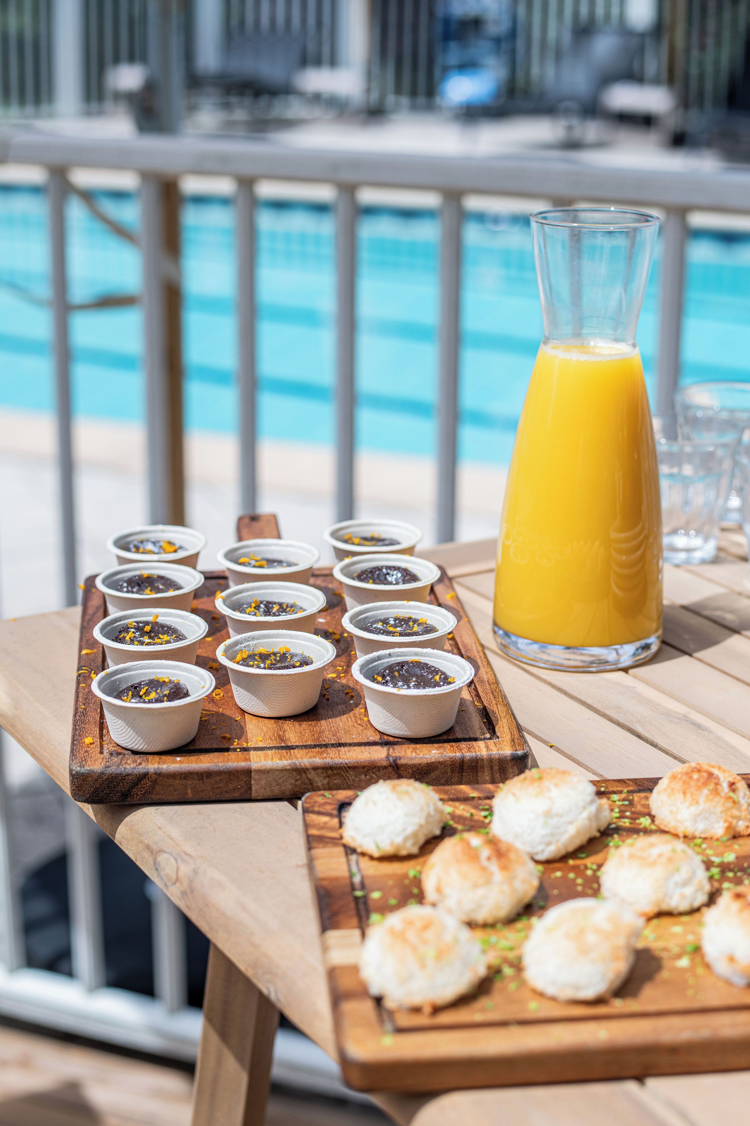 A wooden table with food items and a glass of orange juice by a poolside.