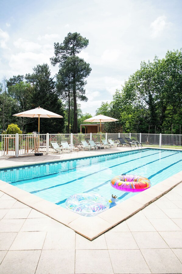 Una piscina con sillas de descanso y sombrillas, rodeada por una valla blanca y árboles.