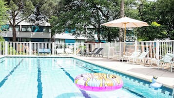 Una piscina con sillas de descanso y sombrillas, rodeada por una valla blanca y árboles.