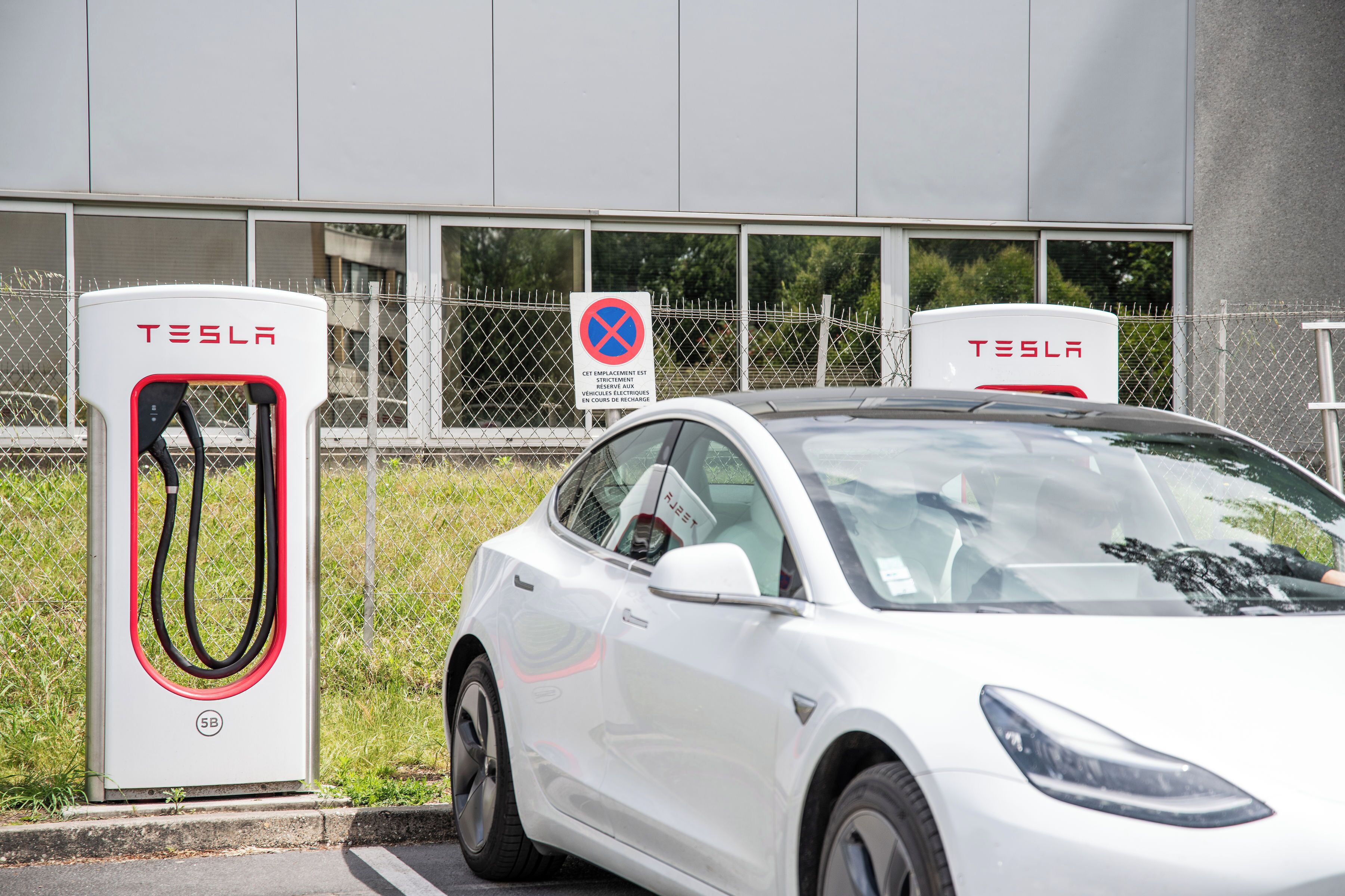 Una estación de carga para Tesla blanca, con una valla y un edificio al fondo.