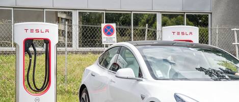 Una estación de carga para Tesla blanca, con una valla y un edificio al fondo.