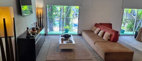 A modern living room with a sofa, a coffee table, a flat-screen TV, and a view of greenery through large windows.