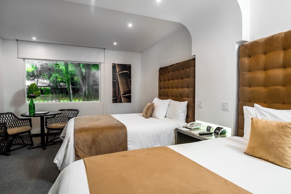 A hotel room with two beds, a small table with chairs, a window with a view of trees, and a wall-mounted telephone.