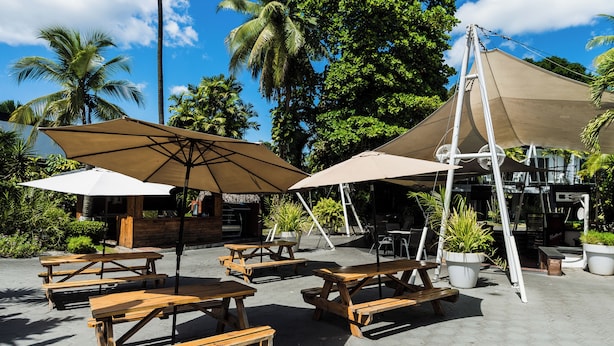Un patio al aire libre con mesas de madera y sombrillas color beige, rodeado de exuberante follaje y palmeras.