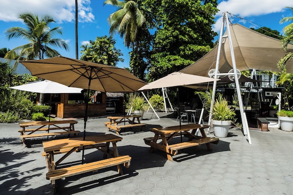 An outdoor patio with wooden tables and beige umbrellas, surrounded by lush greenery and palm trees. - Riande Aeropuerto Hotel & Casino (Tocumen)