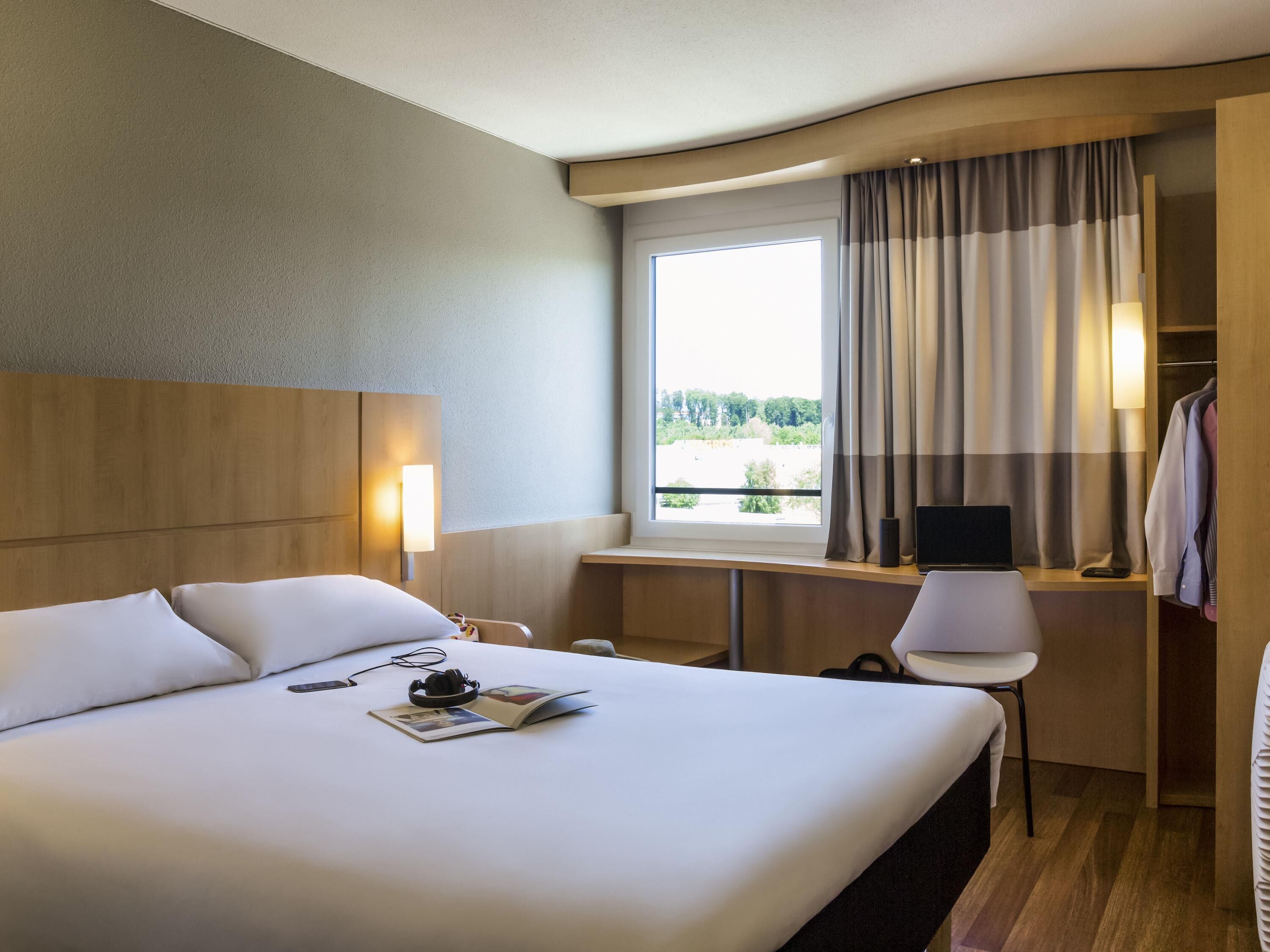 Una habitación de hotel con una cama grande, un escritorio con una silla, una computadora portátil y un ventanal con cortinas.