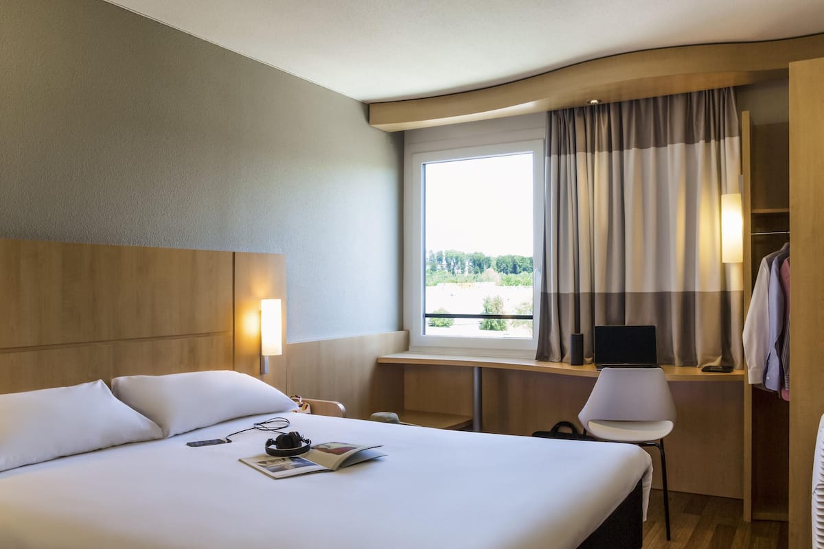 Una habitación de hotel con una cama grande, un escritorio con una silla, una computadora portátil y un ventanal con cortinas.