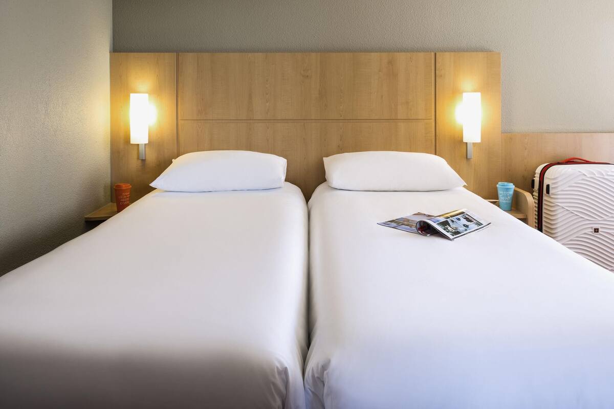 Habitación de hotel con dos camas individuales, cabecero de madera, lámparas de noche, una taza de café roja y una maleta blanca.