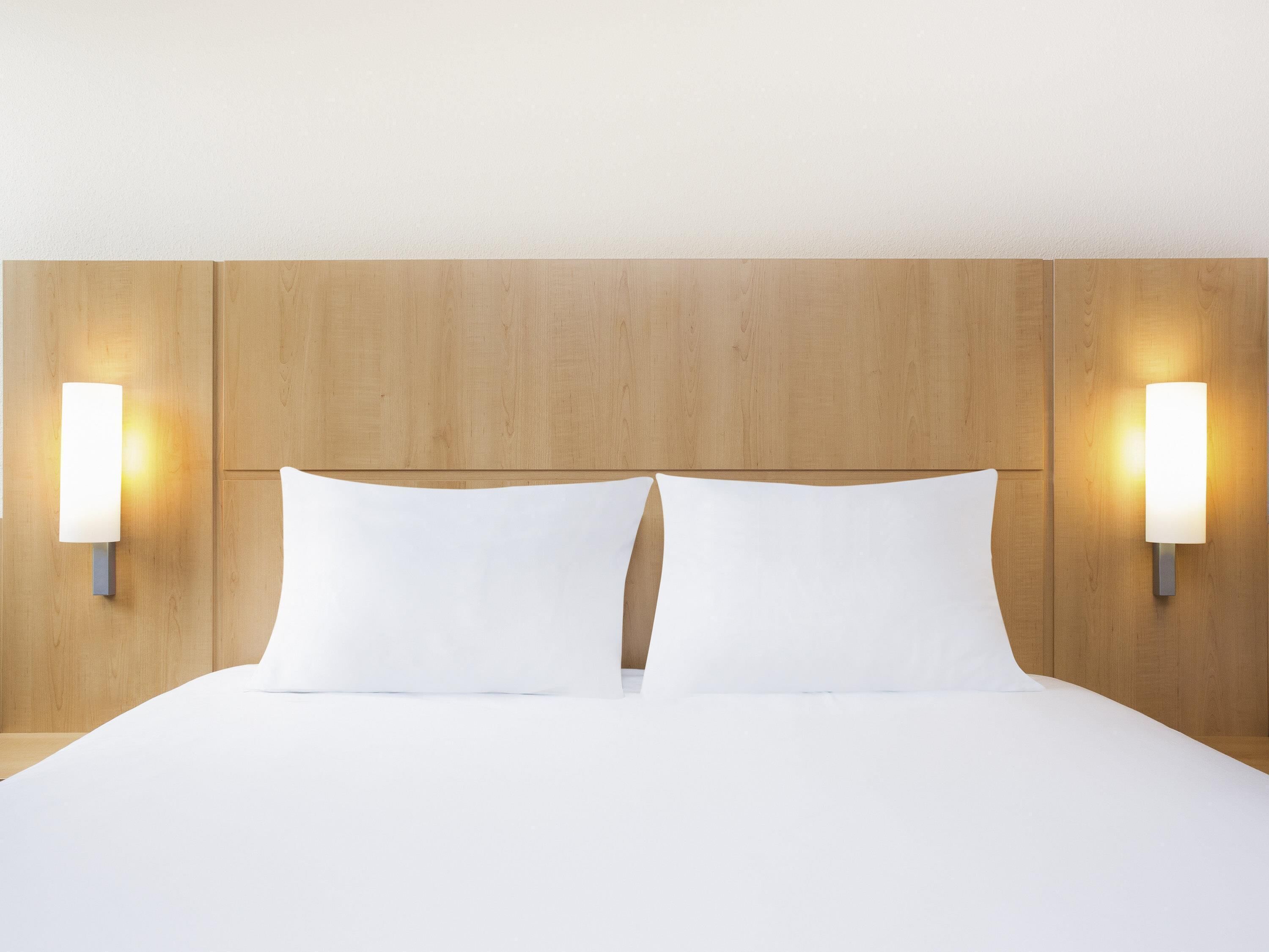 Una habitación de hotel con cabecera de madera, dos almohadas blancas e iluminación empotrada en la pared.