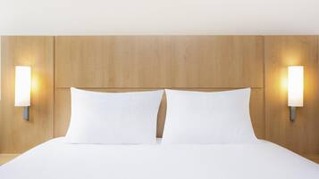 Una habitación de hotel con cabecera de madera, dos almohadas blancas e iluminación empotrada en la pared.