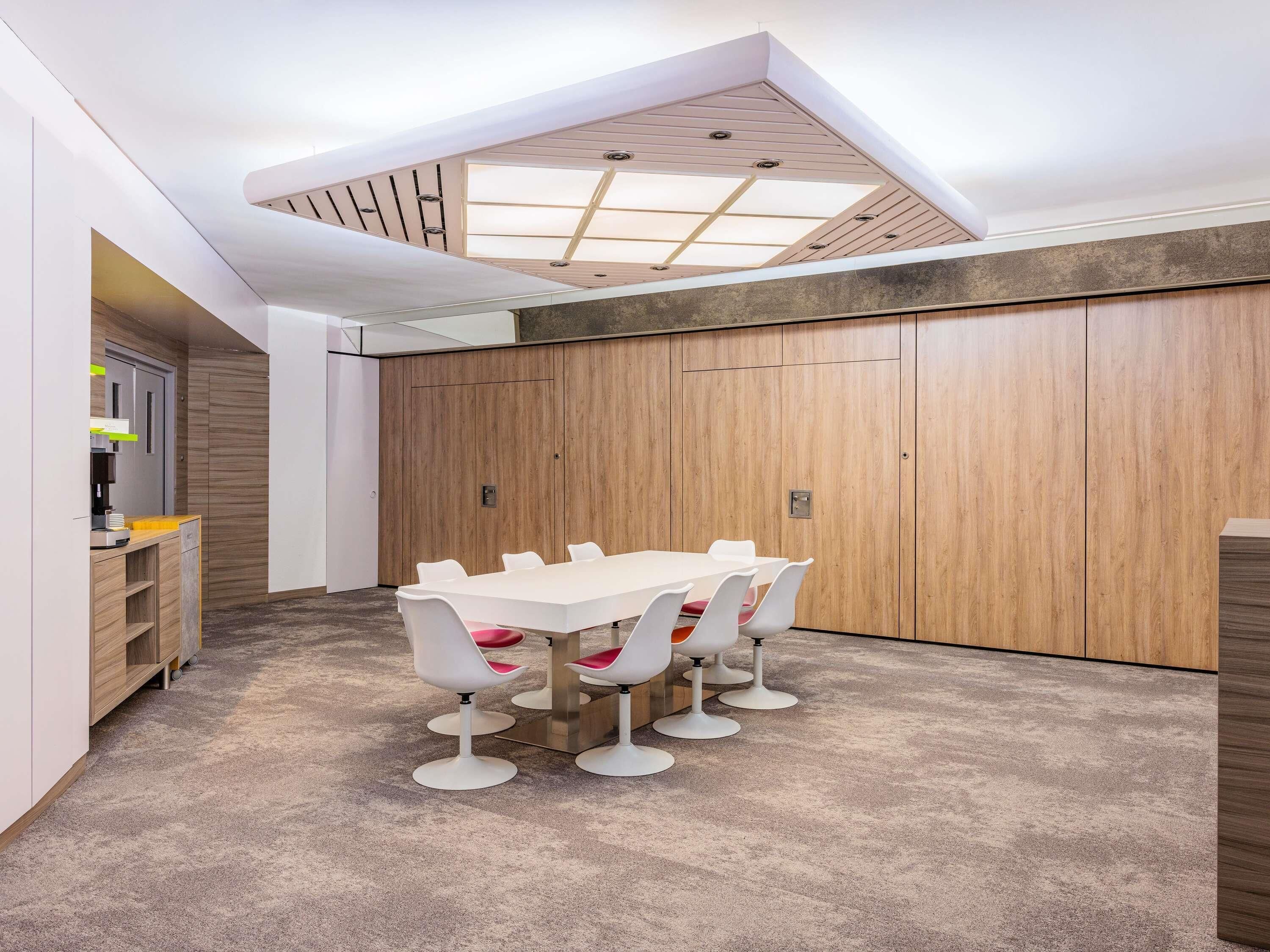 Ein Konferenzraum mit langem Tisch und weißen Stühlen, einer Holzwand als Raumteiler und einer Decke mit Oberlichtern.