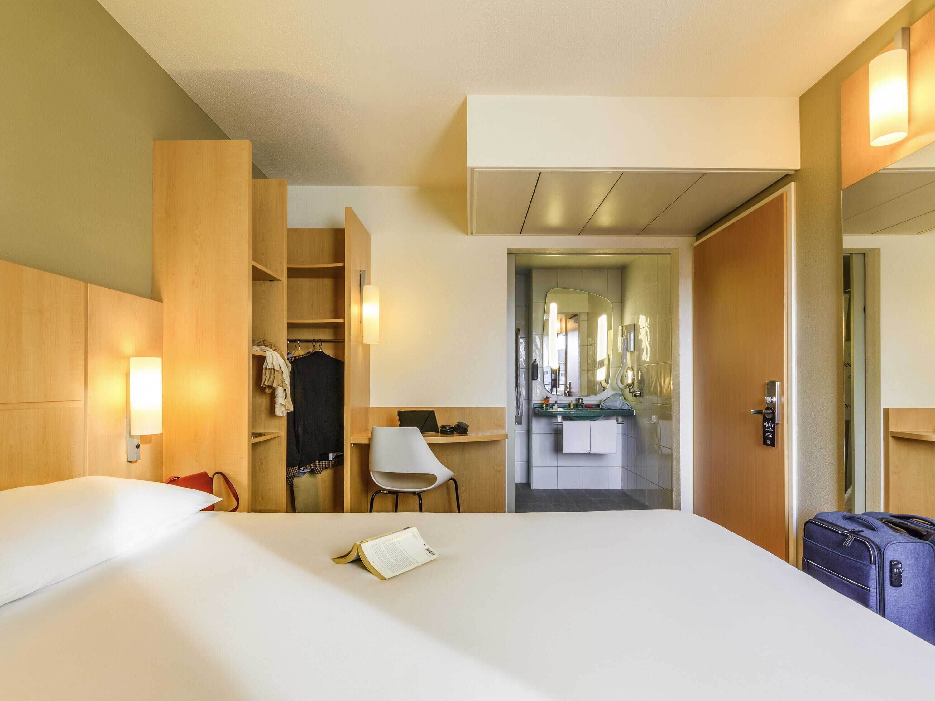 A hotel room with a bed, a desk, a chair, a wardrobe, a bathroom with a mirror, and a suitcase.