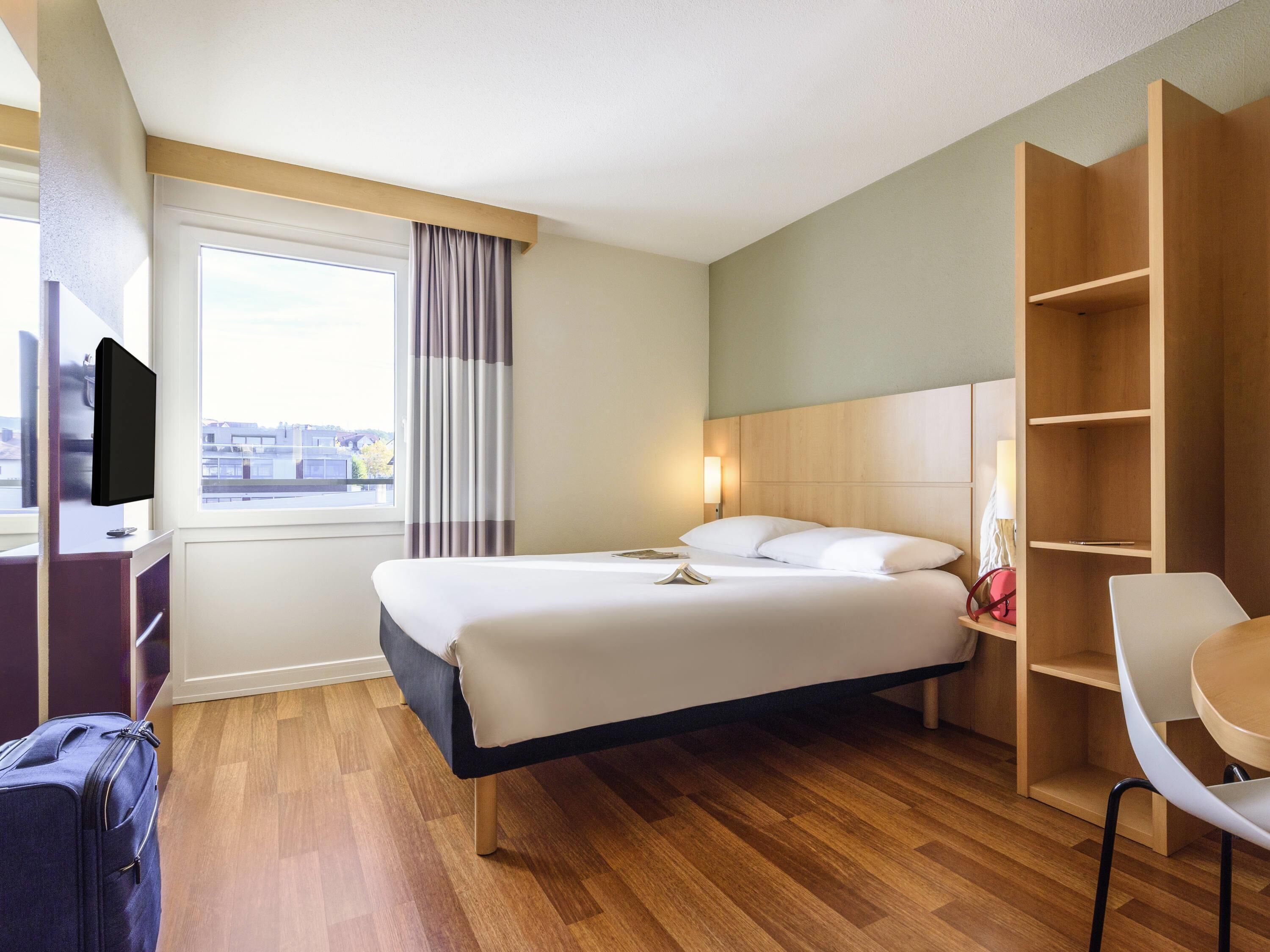 Ett hotellrum med en säng, ett sängbord, en lampa, en garderob, ett skrivbord, en stol och en tv.