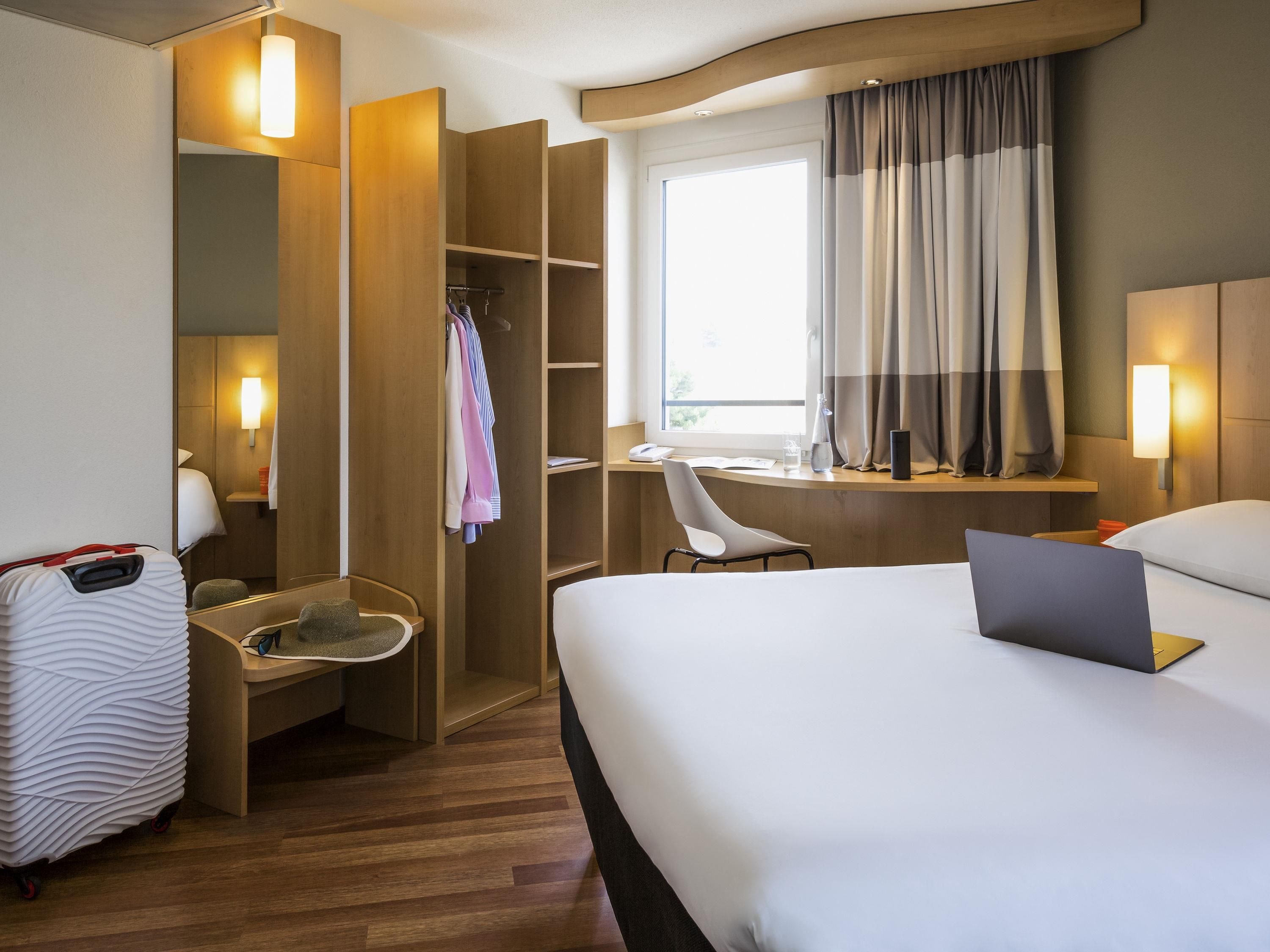 A hotel room with a bed, a desk, a chair, a wardrobe, and a suitcase.