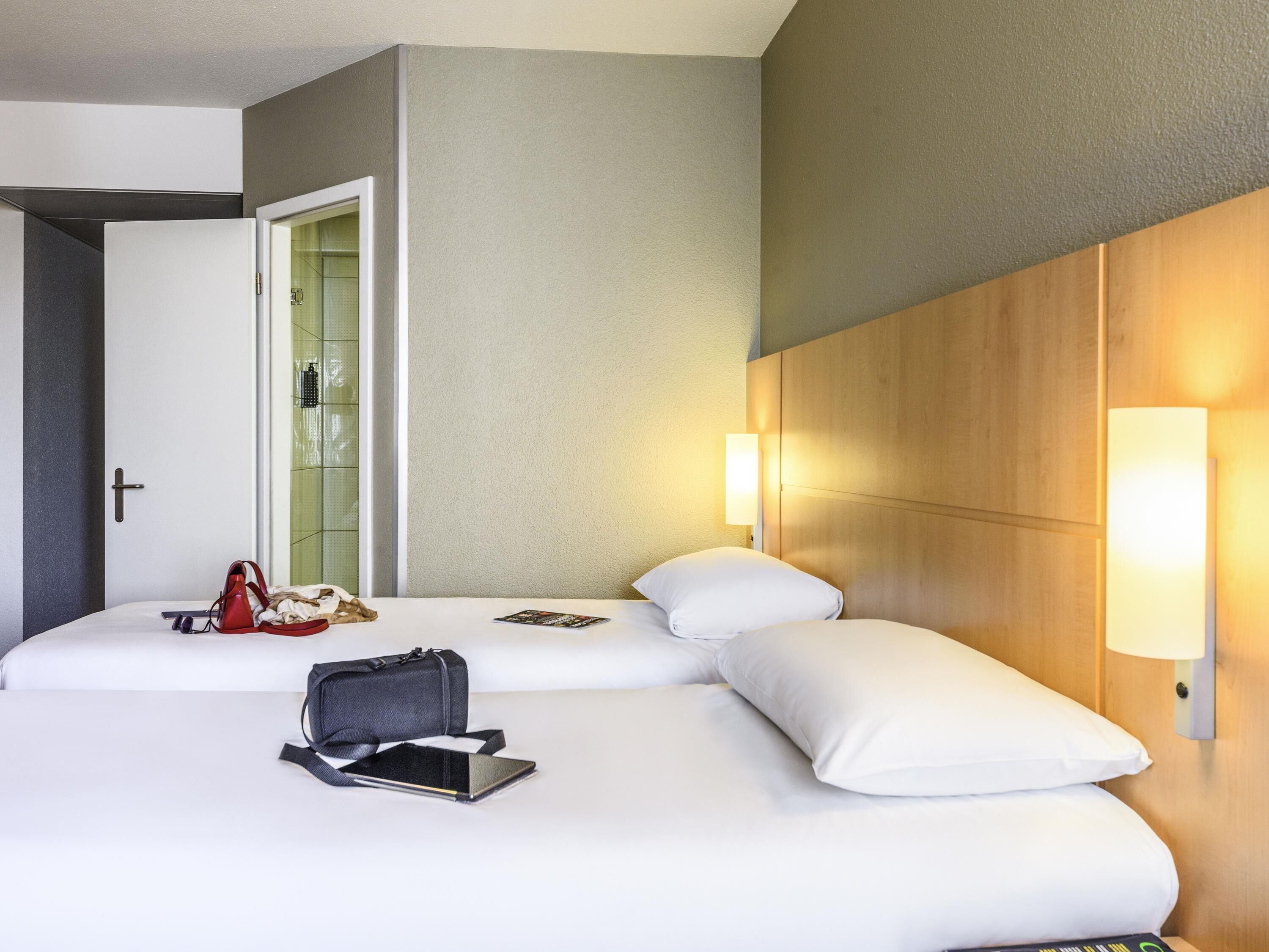 A hotel room with two beds, a door, a wall-mounted light, and a bag on the bed.