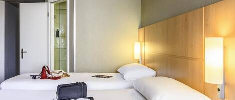 A hotel room with two beds, a door, a wall-mounted light, and a bag on the bed.
