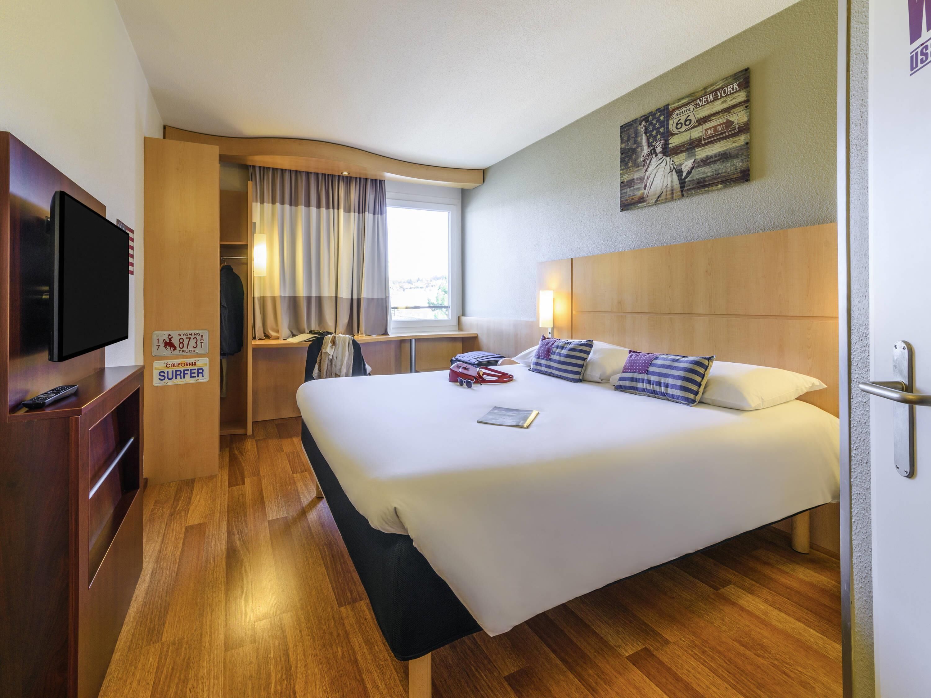 Quarto de hotel com uma cama grande, televisão, escrivaninha com cadeira, janela com cortinas e uma obra de arte na parede.