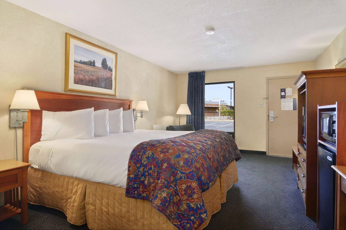 A hotel room with a large bed, two bedside lamps, a wooden headboard, a framed picture on the wall, a desk with a lamp, a dresser with a TV, and a door leading to a parking area.